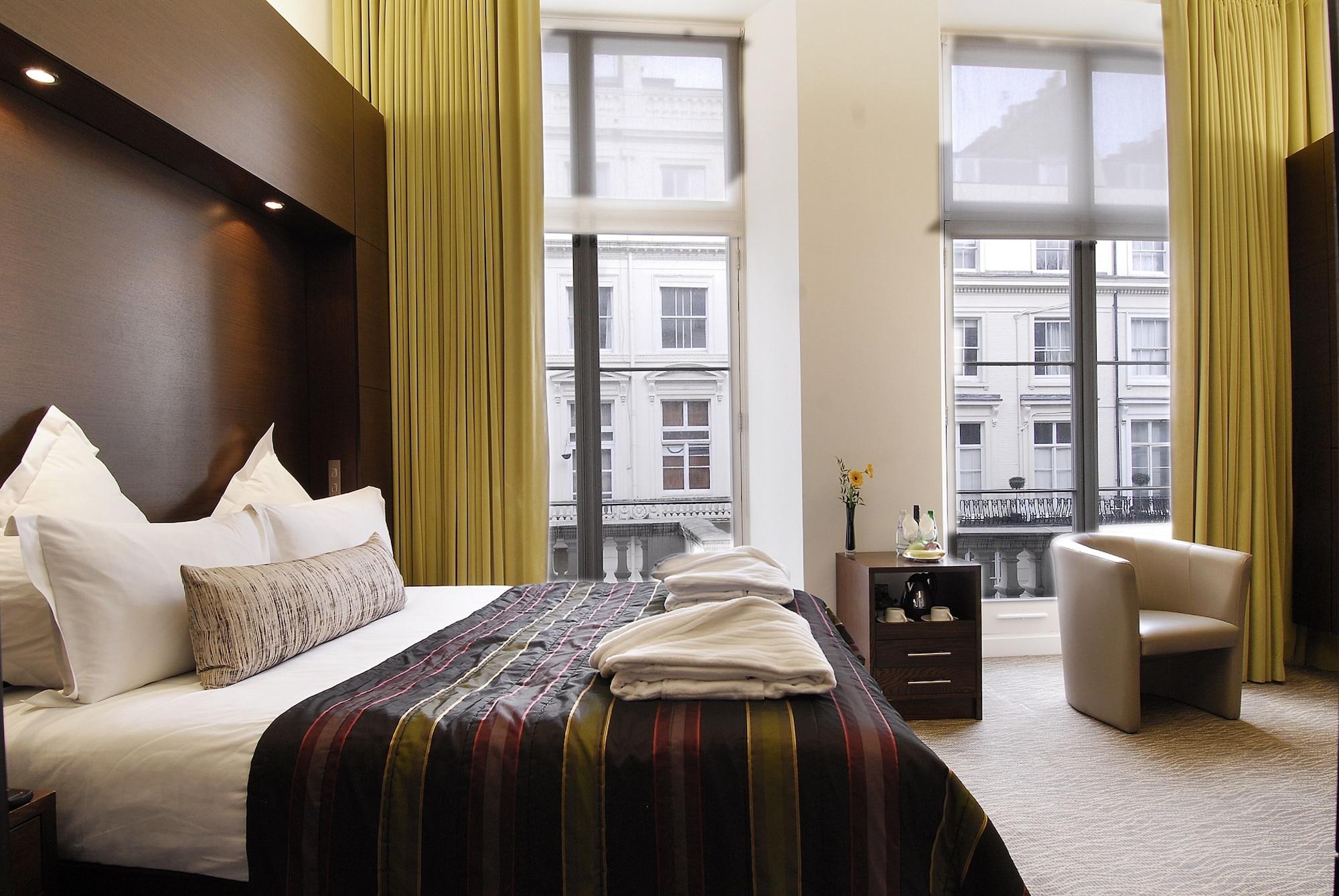 Inhabit Queen'S Gardens, A Member Of Design Hotels Londres Exterior foto The photo shows a modern hotel room with a large bed dressed in white linens and a patterned throw. There are two towels neatly stacked at the foot of the bed. The walls are decorated in neutral tones, and the room features large windows with sheer a