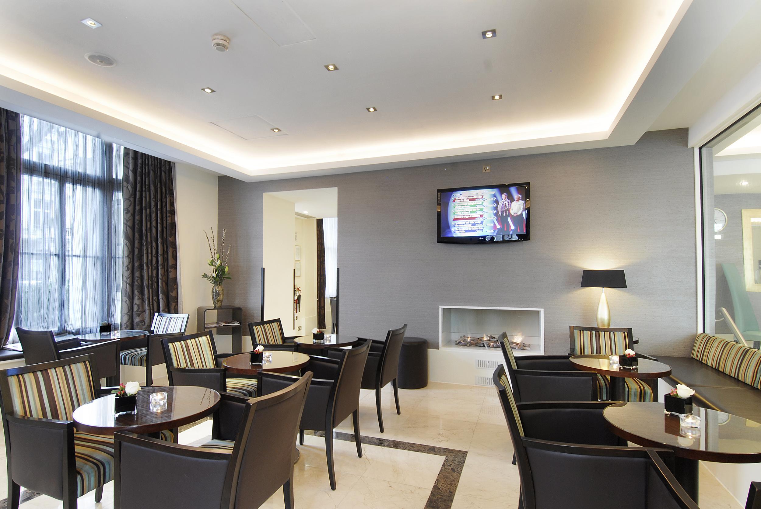 Inhabit Queen'S Gardens, A Member Of Design Hotels Londres Exterior foto The photo shows an elegant interior of a lounge or café area. It features several tables with dark wooden surfaces surrounded by chairs. The walls are adorned in neutral tones, with a soft ceiling light illuminating the space. Large windows allow nat